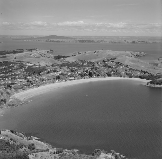 Aerial photograph of the curve of Oneroa Beach and the houses behind it, with Rangitoto Island on the horizon